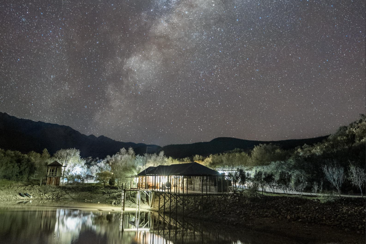 A scenic night view of a stilt-supported structure situated on a waterfront, featuring a raised deck and illuminated exterior under a starry sky.