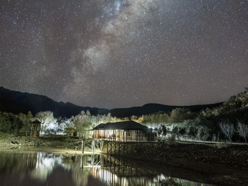 A scenic night view of a stilt-supported structure situated on a waterfront, featuring a raised deck and illuminated exterior under a starry sky.