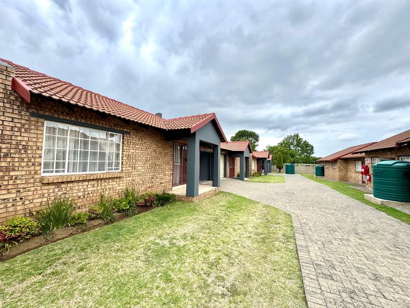 Exterior view of a townhouse complex featuring face brick facades, tiled roofs, paved driveways, and established garden areas with water storage tanks.