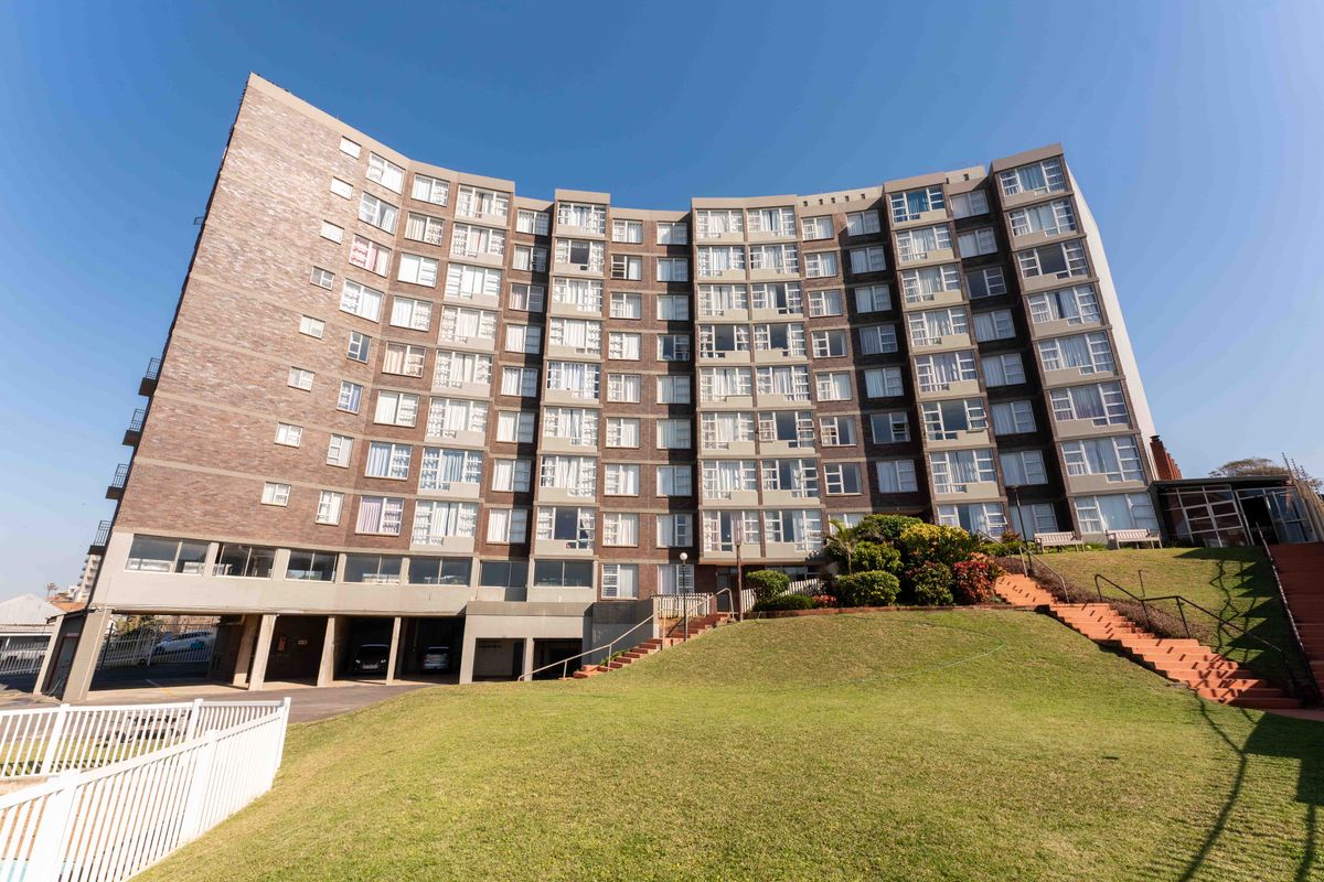 A wide-angle view of the rear elevation of a multi-story brick apartment building featuring landscaped grounds, concrete staircases, and ground-level undercover parking.
