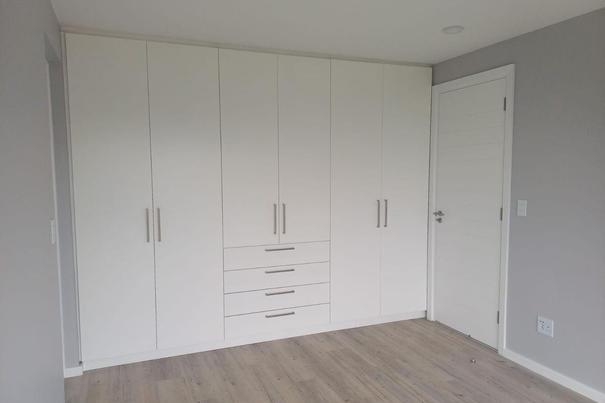 A clean, modern bedroom featuring extensive floor-to-ceiling built-in cupboards, light wood-look laminate flooring, and neutral grey walls.
