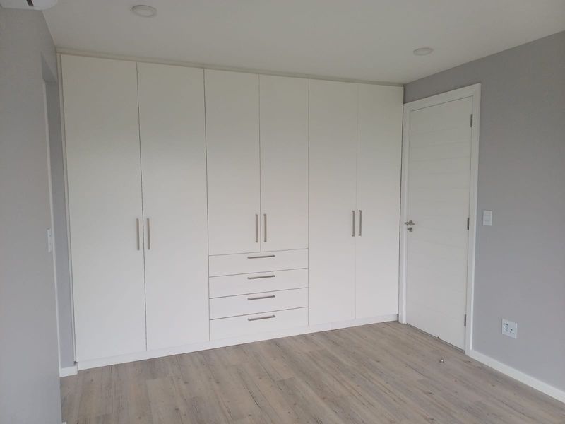 A clean, modern bedroom featuring extensive floor-to-ceiling built-in cupboards, light wood-look laminate flooring, and neutral grey walls.