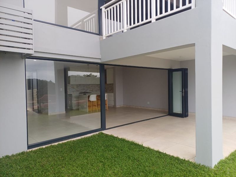 A modern covered patio area featuring large stacking glass doors that open into the interior living space, with tiled flooring and a balcony above.