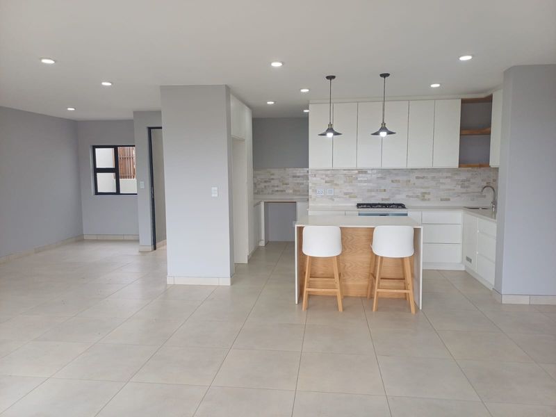 Modern open-plan kitchen and living area featuring sleek white cabinetry, a central island, stone tile flooring, and contemporary recessed and pendant lighting.