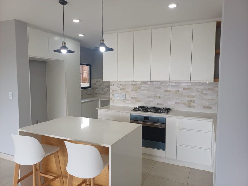 Modern kitchen featuring white handleless cabinetry, stone countertops, a central island, gas hob, built-in oven, and a decorative stone tile backsplash.