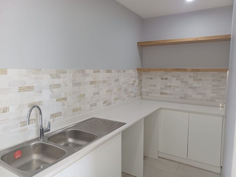 A clean, modern scullery featuring a double stainless steel sink, stone countertops, tiled backsplash, and ample built-in cabinetry with floating timber shelving.