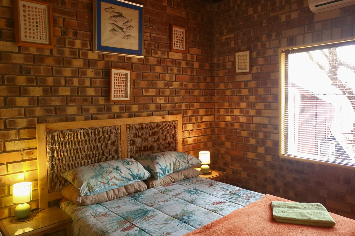 A cozy bedroom featuring rustic exposed brick walls, a wall-mounted air conditioning unit, and natural light filtered through venetian blinds.