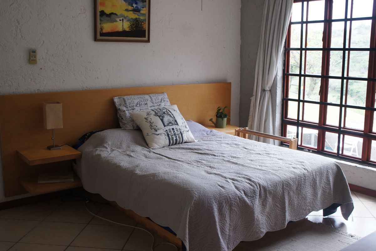 A bedroom featuring a built-in wooden headboard with integrated floating bedside tables, tiled flooring, and a large grid-pattern window providing natural light.
