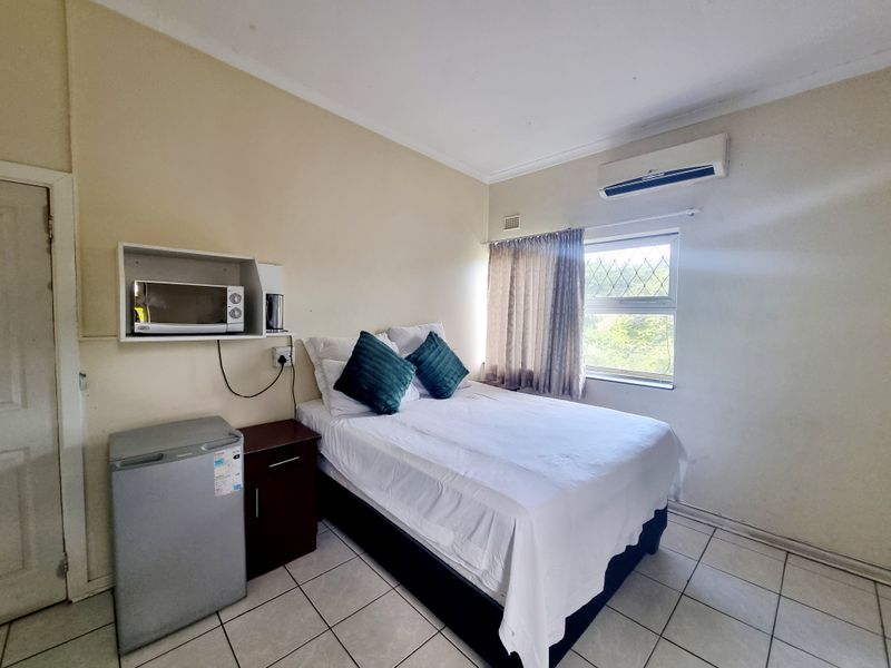 A functional bedroom featuring tiled flooring, a wall-mounted air conditioning unit, and a window with security grilles.