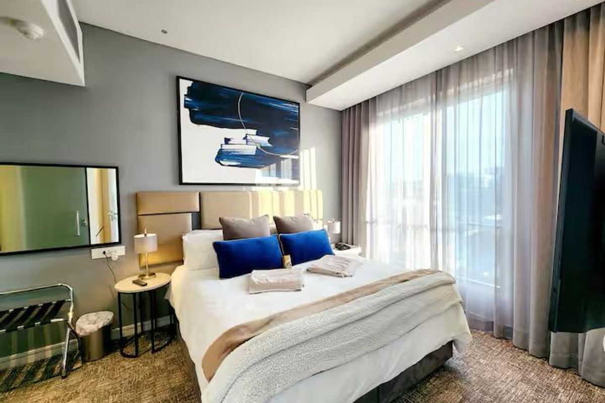 A well-maintained bedroom featuring a built-in headboard, recessed ceiling lighting, and large windows with sheer curtains providing ample natural light.