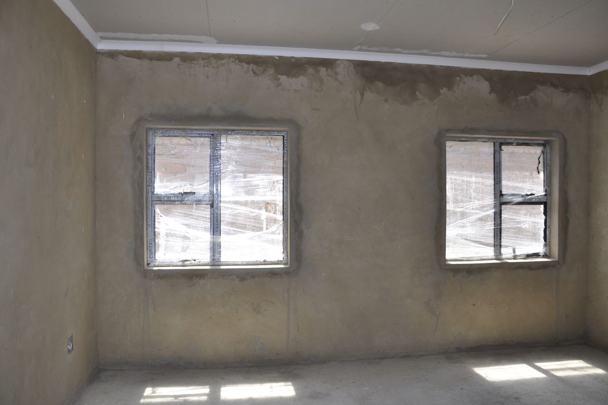 An unfinished bedroom interior featuring raw plaster walls, concrete flooring, and two installed aluminium-framed windows with protective plastic film still attached.