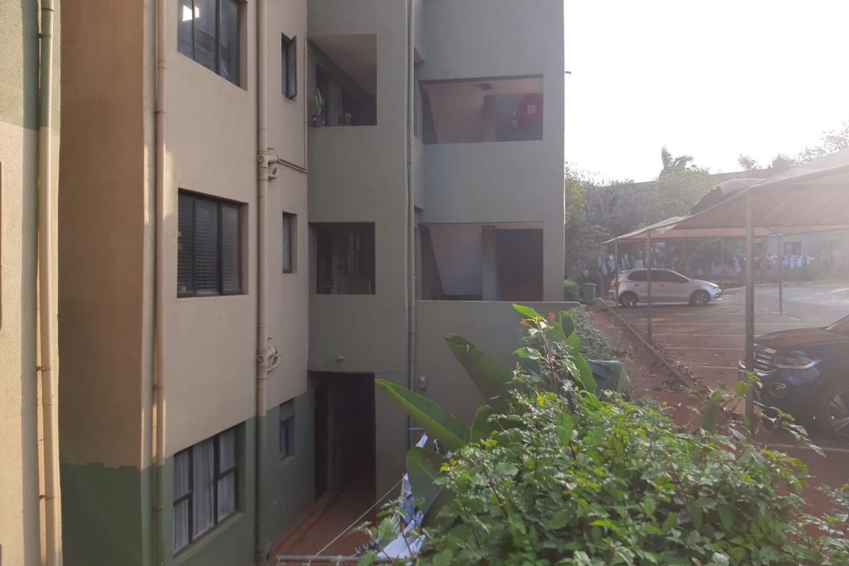 Exterior view of a multi-story apartment building showing the rear facade, external plumbing, and adjacent carport parking area.