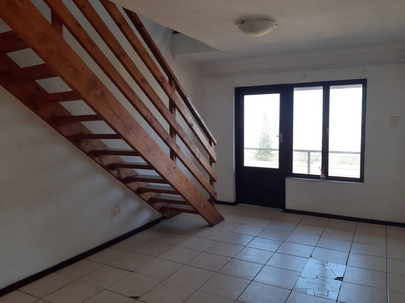 An empty living area featuring a wooden staircase, tiled flooring with visible damage, and access to an outdoor balcony through a glass door.