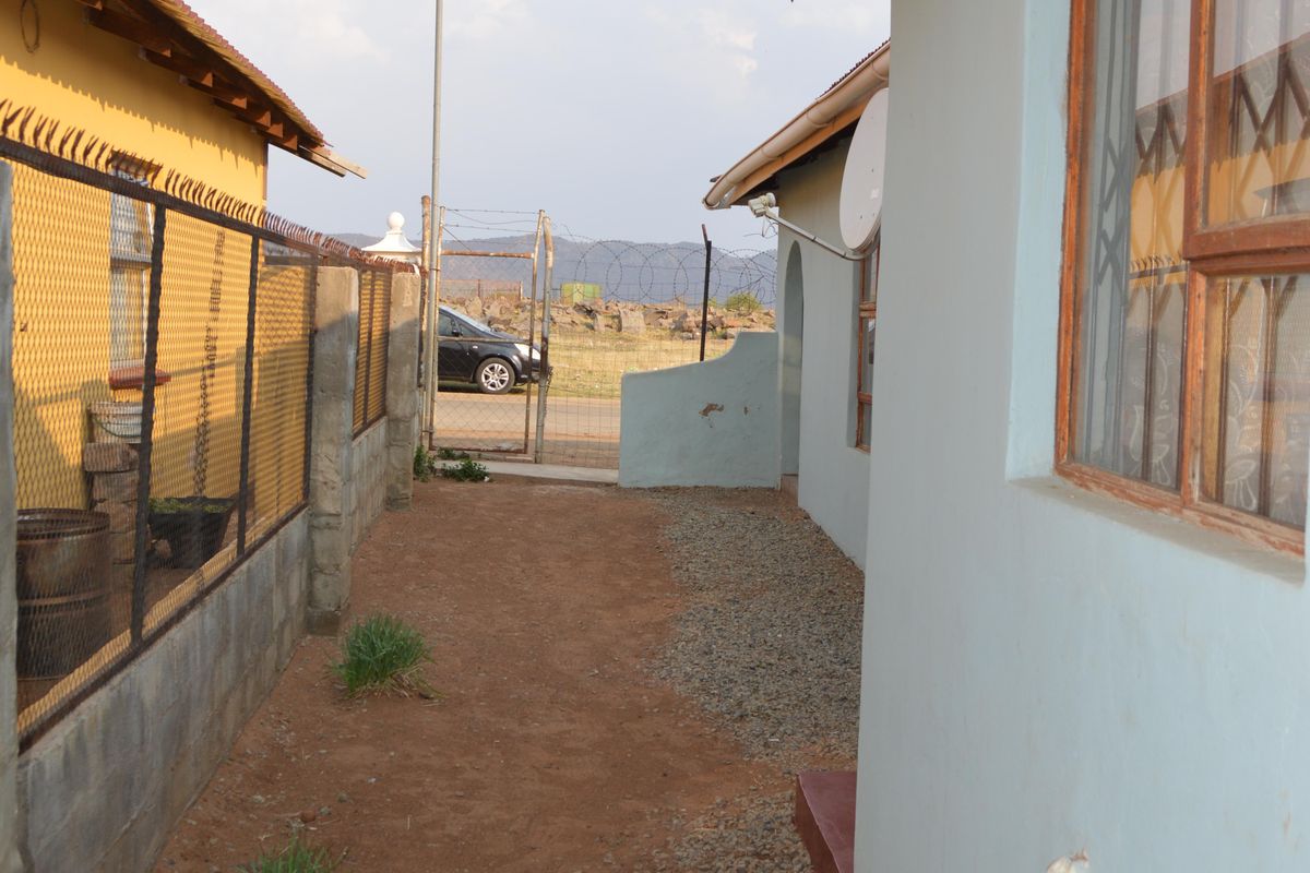 A narrow side passage between two residential buildings featuring gravel and dirt ground, security fencing, and basic exterior wall finishes.