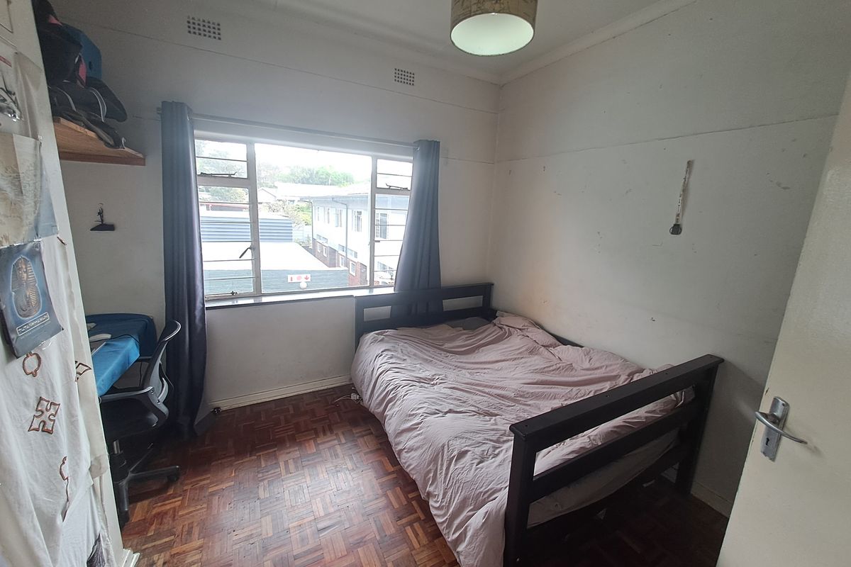 A compact bedroom featuring traditional parquet flooring and a large steel-framed window, offering a functional space in need of cosmetic updates.