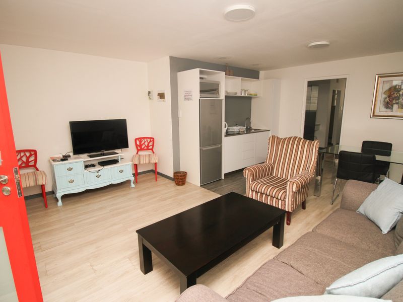 Open-plan living area featuring light laminate flooring, a compact kitchenette with built-in cabinetry, and recessed ceiling lighting.