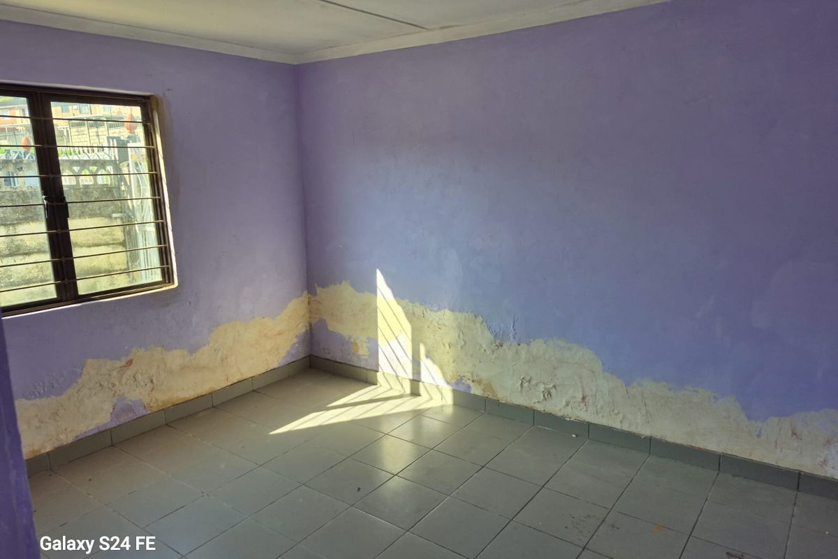 An empty bedroom featuring tiled flooring and security bars on the window, currently showing significant wall plaster damage and peeling paint.