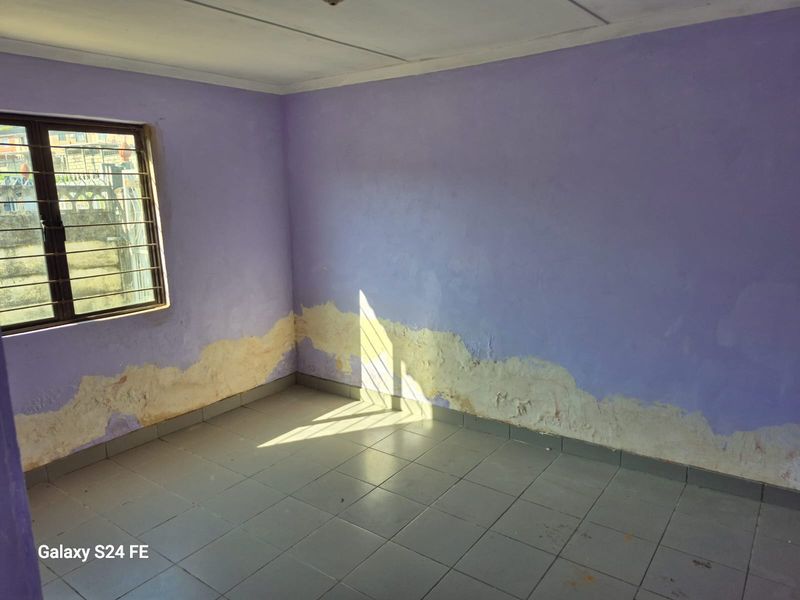 An empty bedroom featuring tiled flooring and security bars on the window, currently showing significant wall plaster damage and peeling paint.