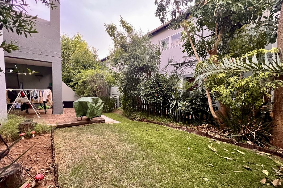 A private rear garden featuring a paved patio area, established trees, and a lawn, providing a quiet outdoor space for the apartment.