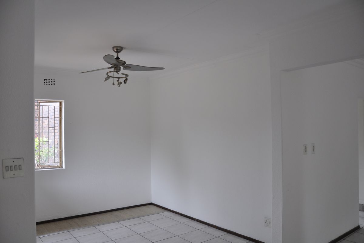 A bright, empty living area featuring neutral tiled flooring, a ceiling fan, and a security-grilled window, offering a clean canvas for personalization.