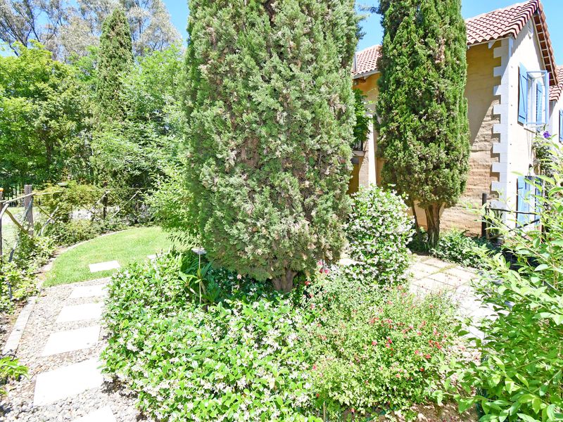 A lush, well-maintained garden area featuring mature cypress trees, stone pathways, and a view of the home's exterior facade with classic shutters.