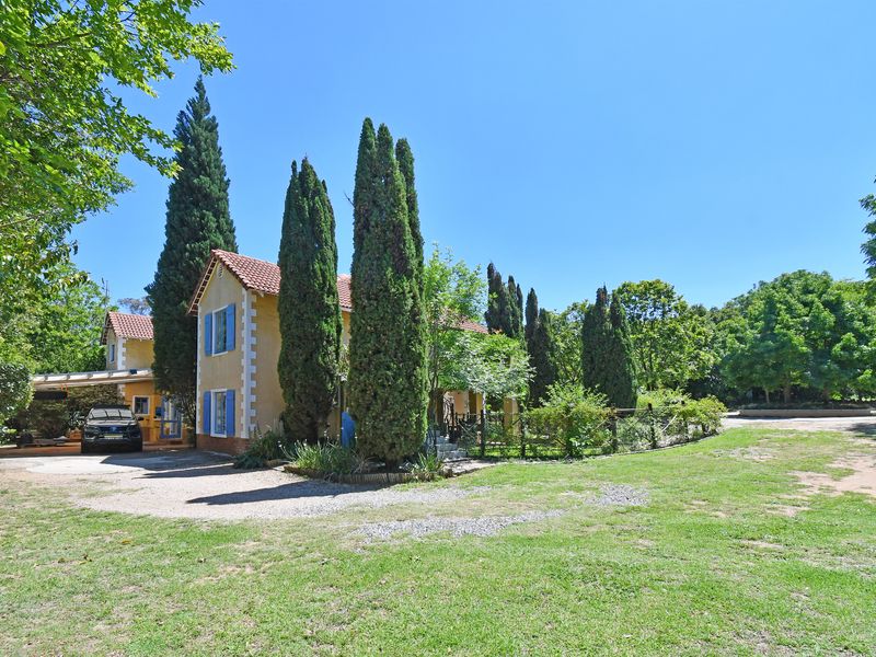 A charming two-story house featuring a terracotta tiled roof, stucco exterior, and blue shutters, set within a spacious, well-maintained garden with a carport.