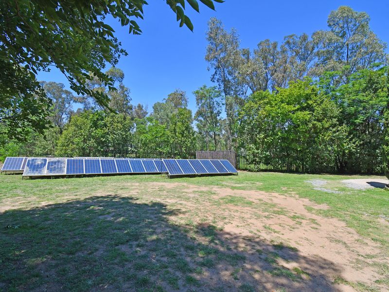 A spacious garden area featuring a large ground-mounted solar panel array set against a backdrop of mature trees and perimeter fencing.