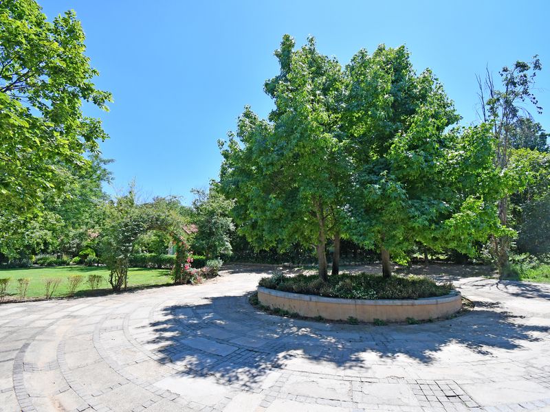 A spacious garden area featuring a circular stone-paved driveway, a central raised garden bed with mature trees, and a charming rose archway.