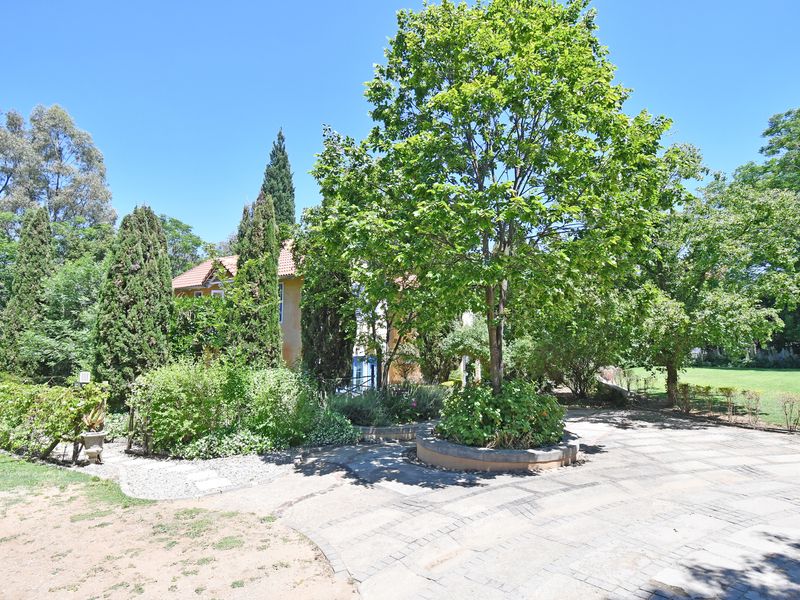 A spacious, well-maintained garden area featuring mature trees, established landscaping, and a paved driveway leading toward the property.