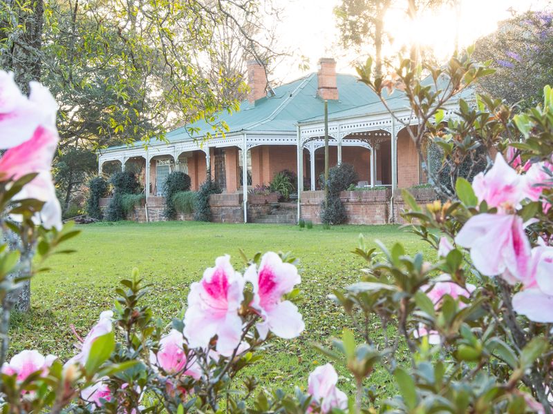 A charming historic brick home featuring a wide covered veranda with ornate fretwork, set within a lush garden with mature trees.