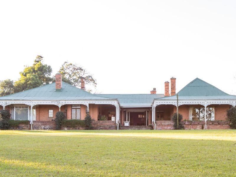 A spacious rear view of a traditional brick house featuring a wide veranda with decorative ironwork, metal roofing, and extensive surrounding lawn.
