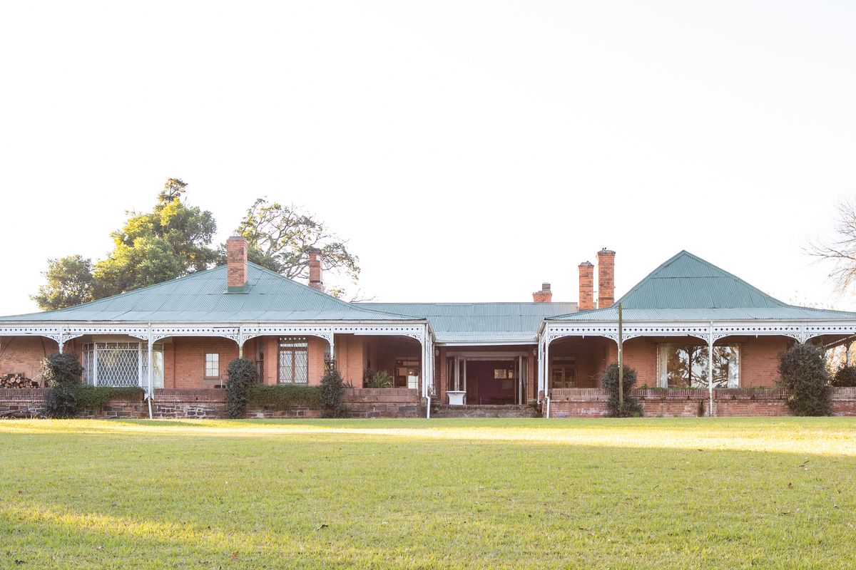 A spacious rear view of a traditional brick house featuring a wide veranda with decorative ironwork, metal roofing, and extensive surrounding lawn.