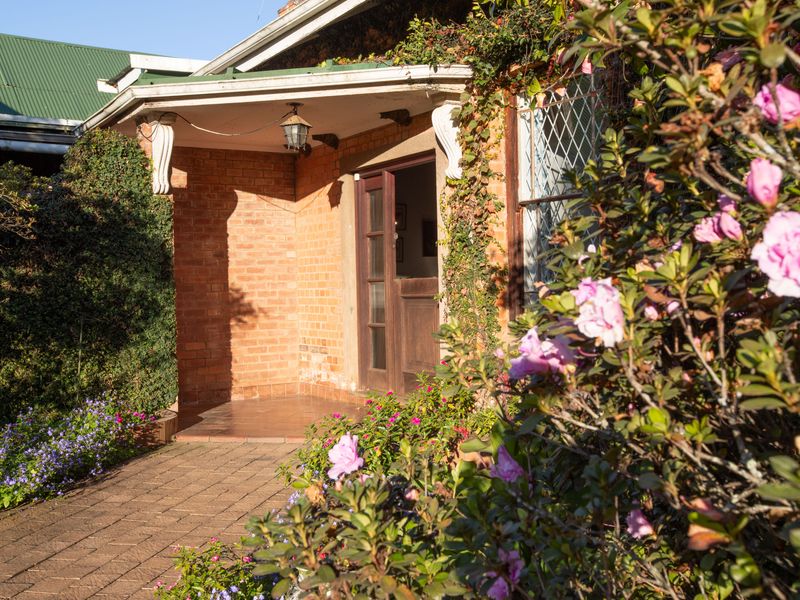 A charming brick-faced front entrance featuring a covered porch, leaded glass windows, and a paved walkway surrounded by established garden landscaping.