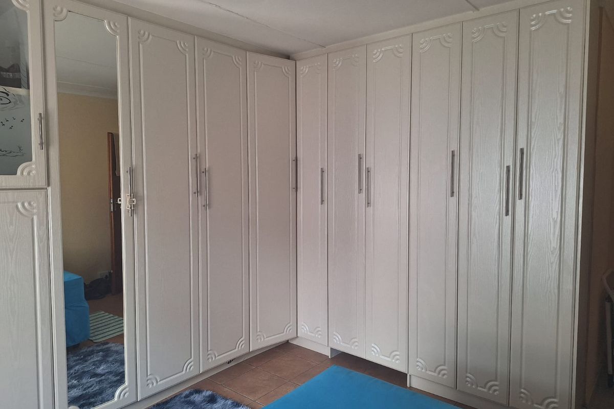 A bedroom featuring extensive white built-in cupboards with decorative detailing and a full-length mirrored door, set against tiled flooring.