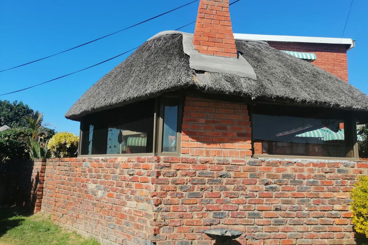 Exterior view of a brick structure featuring a thatched roof, brick chimney, and aluminium window frames, showing signs of wear on the roof and masonry.