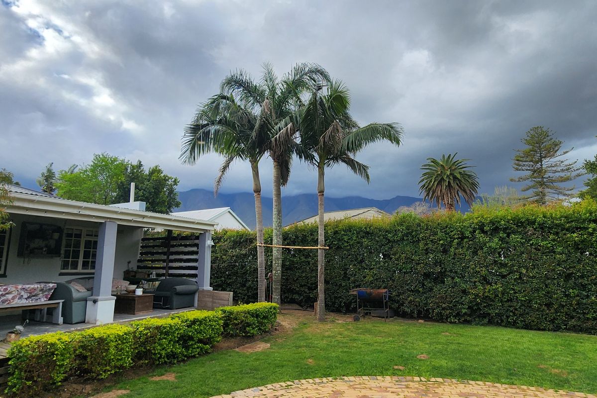 A well-maintained rear garden featuring a covered patio, brick paving, lush lawn, and mature hedging with scenic mountain views in the background.