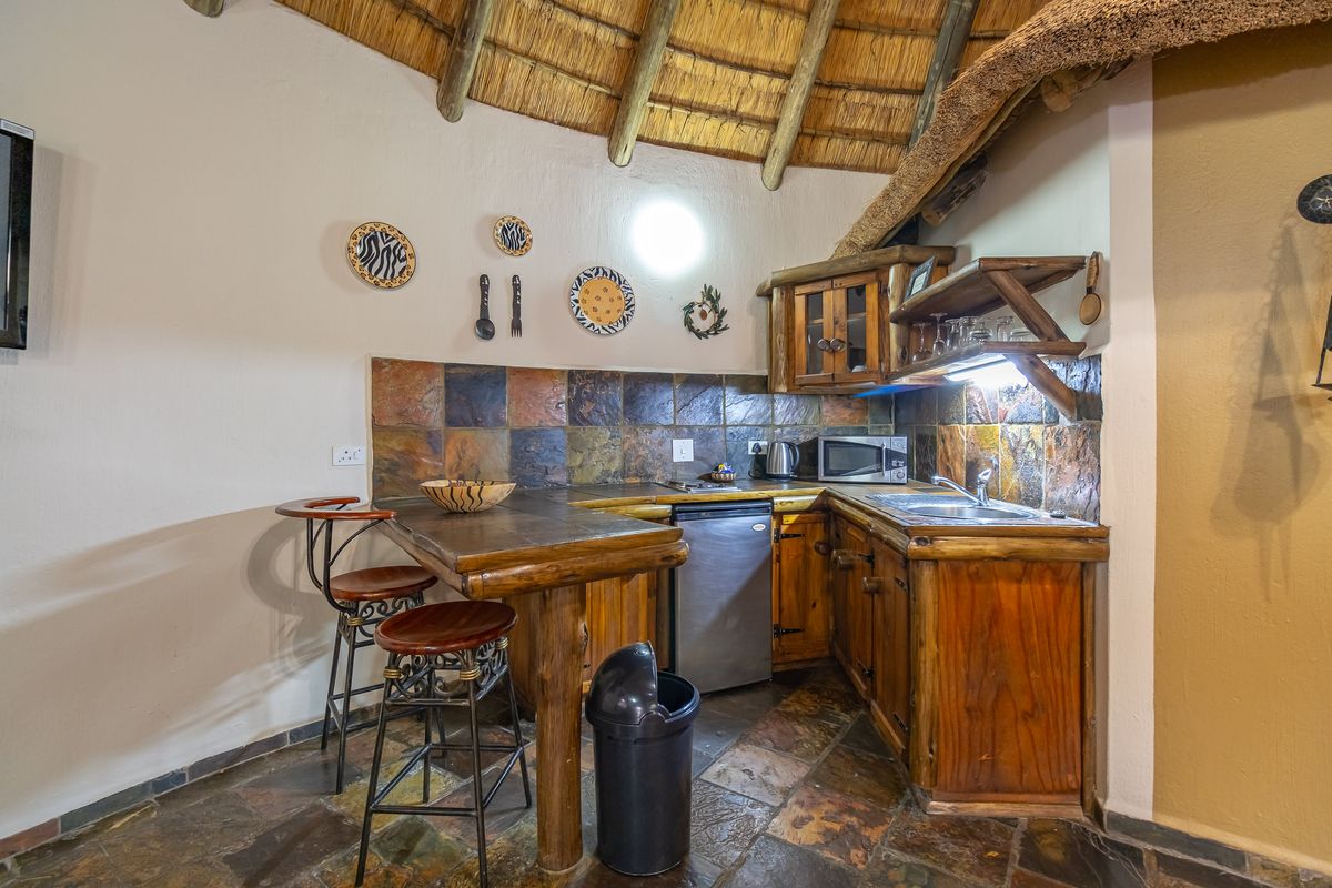Compact rustic kitchen featuring slate tile flooring, a slate backsplash, wooden cabinetry, and a built-in breakfast bar under a traditional thatched roof.