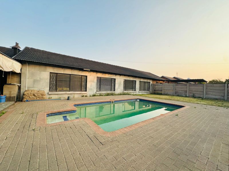 A rear exterior view featuring a swimming pool with brick paving, concrete boundary wall, and a single-story house with tiled roof and security bars.
