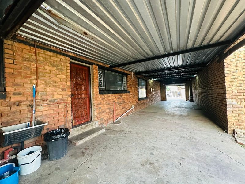 A covered carport area featuring exposed brick walls, a corrugated metal roof, and a utility sink, providing functional outdoor vehicle storage or workspace.