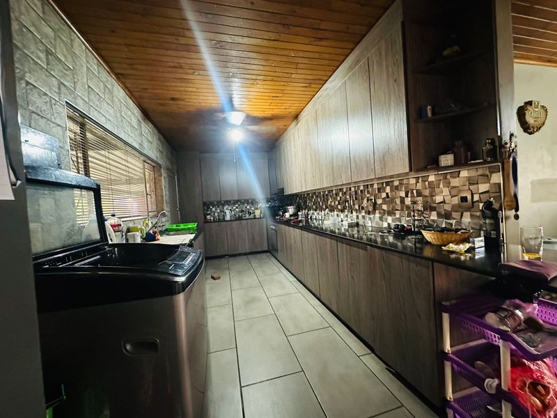 A galley-style kitchen featuring wood-look cabinetry, dark countertops, a mosaic tile backsplash, and a timber-paneled ceiling with tiled flooring.