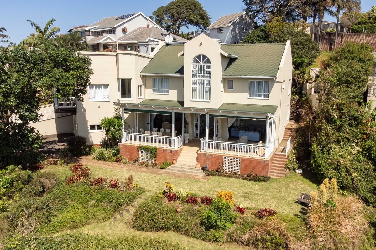 Aerial view of the rear elevation featuring a multi-level home with a covered patio, brick foundation, and established garden.