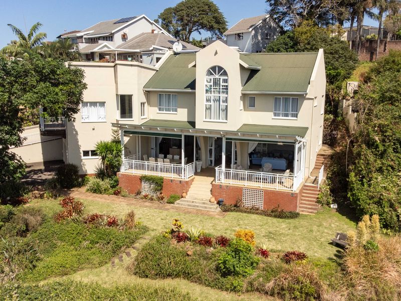 Aerial view of the rear elevation featuring a multi-level home with a covered patio, brick foundation, and established garden.