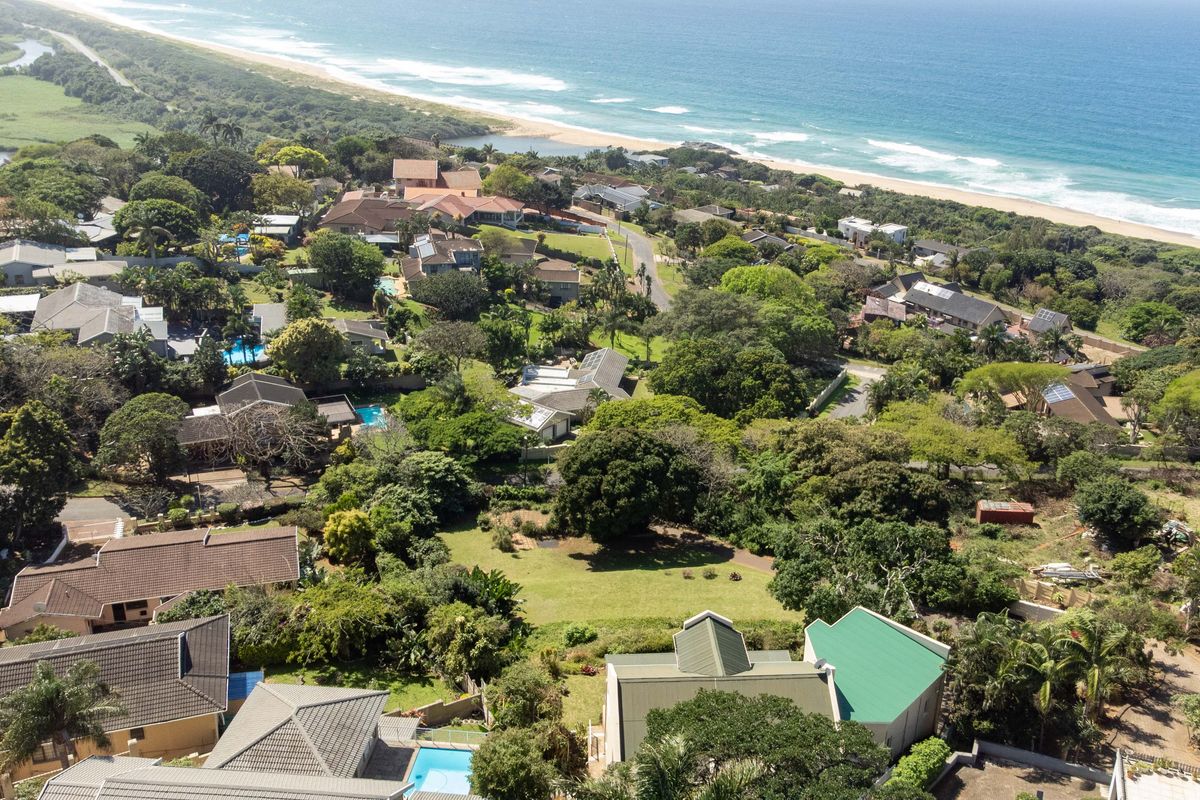 An aerial view of a coastal residential estate featuring lush greenery, multiple homes with tiled roofs, and proximity to the beach.