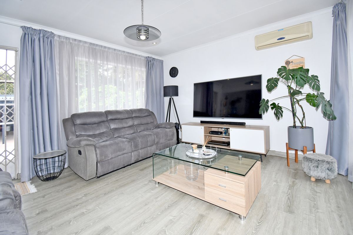 A well-maintained living room featuring light-toned laminate flooring, a wall-mounted air conditioning unit, and a sliding glass door with security gate access.
