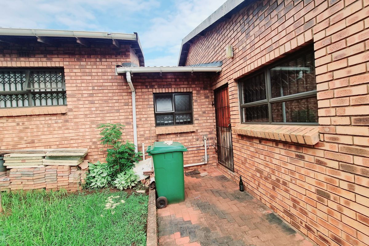 A rear exterior view of the brick house featuring a paved courtyard area, security window bars, and access to the side of the property.