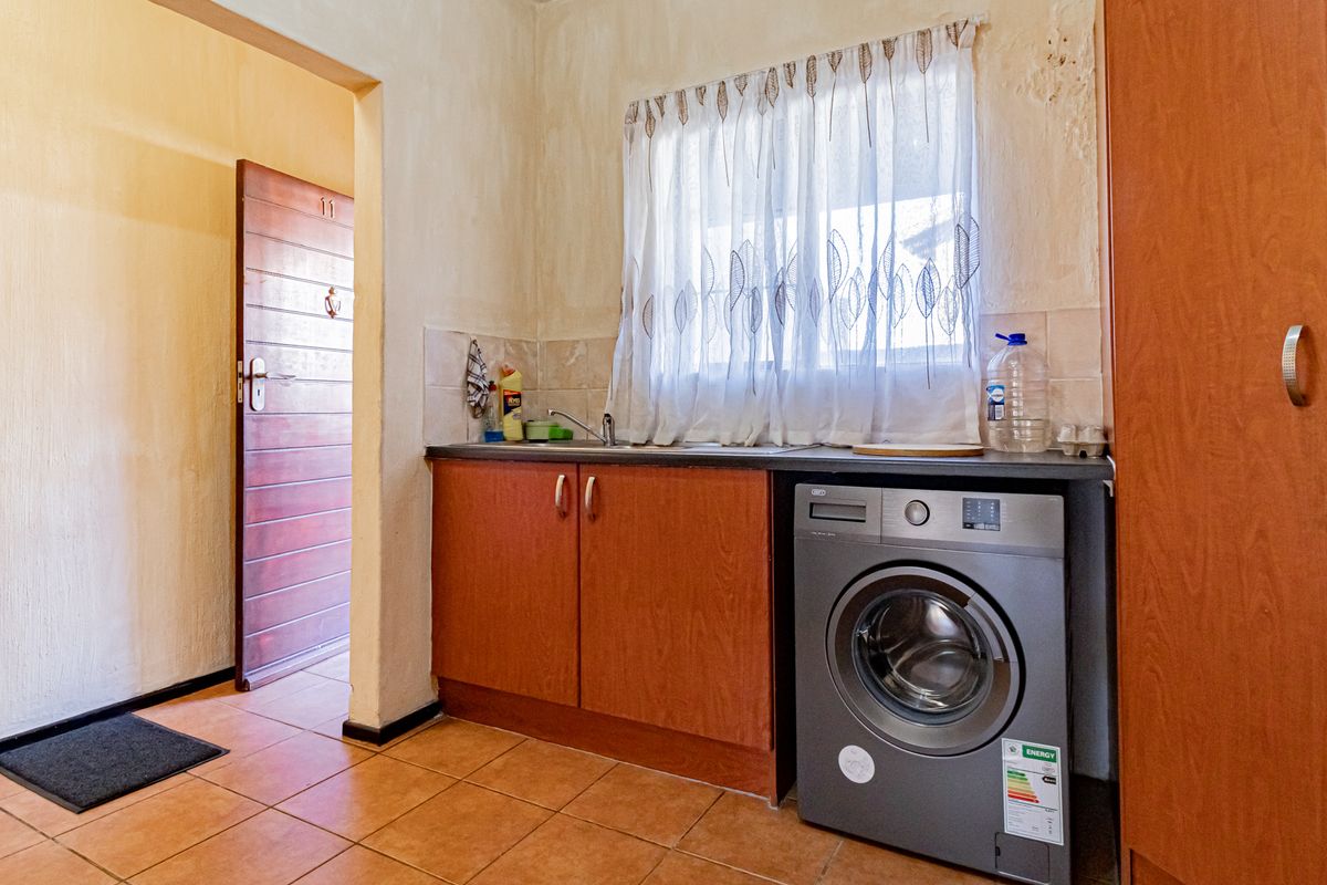 A compact kitchen area featuring wooden cabinetry, a built-in sink, and space for a washing machine, with tiled flooring throughout.