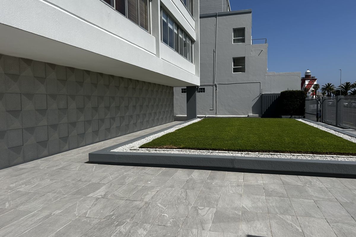 A well-maintained rear exterior area featuring a tiled patio, a raised artificial lawn with gravel borders, and modern textured wall cladding.