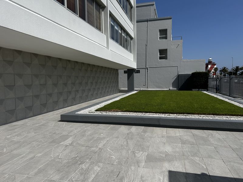 A well-maintained rear exterior area featuring a tiled patio, a raised artificial lawn with gravel borders, and modern textured wall cladding.