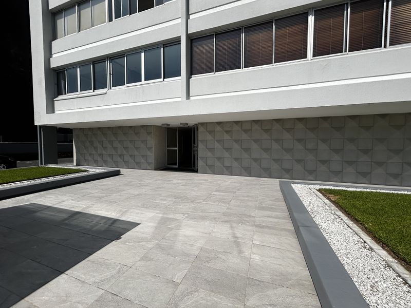 Modern apartment building entrance featuring a paved patio area, textured stone facade detailing, and neatly landscaped garden beds with white gravel borders.