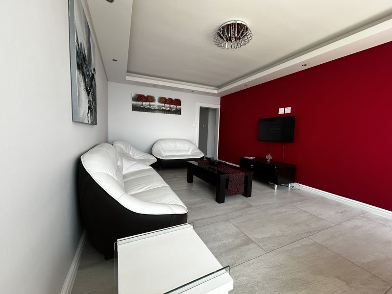 A well-maintained living room featuring large format floor tiles, a recessed ceiling with downlights, and a decorative central chandelier.